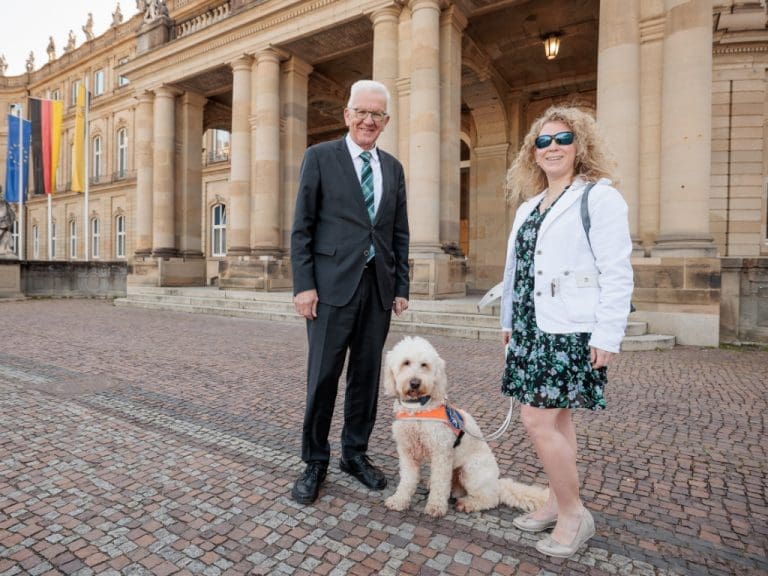 Winfried Kretschmann, der Ministerpräsident von Baden-Württemberg, steht links im Bild. Er lächelt in die Kamera. Er ist Mitte siebzig, hat weiße Haare, trägt eine Brille und einen schwarzen Anzug mit Krawatte. Rechts neben ihm sitzt der Hund Harry, ein Goldendoodle mit blondem, lockigem Fell. Er trägt eine Kenndecke und wird gehalten von Lisa Mümmler, rechts neben ihm. Sie ist eine junge Frau mit blondgelocktem Haar. Sie trägt eine schwarze Sonnenbrille, ein geblümtes Kleid und eine weiße Jacke. Sie lächelt ebenso in die Kamera. Die Gruppe steht vor dem Eingang des Neuen Schlosses in Stuttgart, man sieht Säulen, eine Fassade aus Schilfsandstein, Fenster. Im Hintergrund hängen an drei Masten die Fahnen von Europa, Deutschland und Baden-Württemberg.