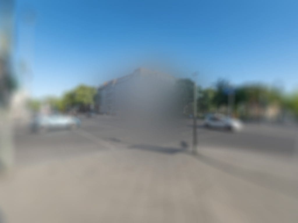 Blick über eine Kreuzung im Sonnenlicht – von einer Straßenecke zur diagonal gegenüberliegenden Ecke. Das Bild ist stark verschwommen, in der Mitte ist nichts zu erkennen, links und rechts davon je ein Auto.