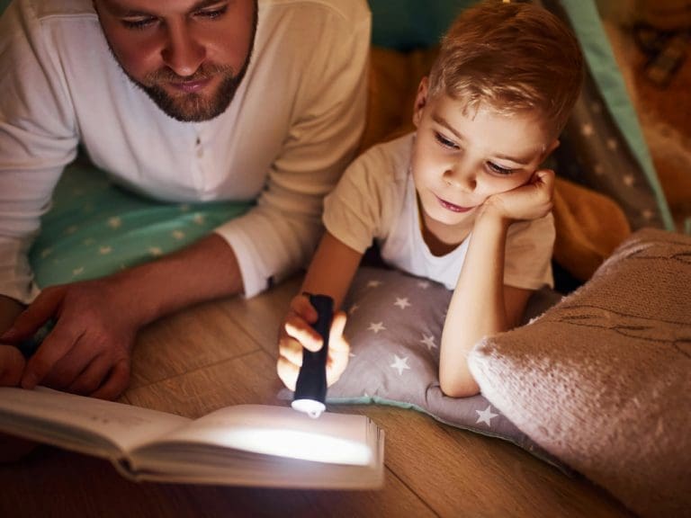 Kind und Vater lesen unter einer Bettdecke Buch mit einer Taschenlampe