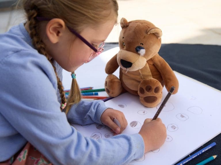 Mädchen mit Brille malt, vor ihr steht ein Stofftier, dem ein Auge fehlt