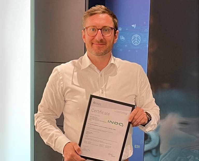 Dr. Matthias Neumann im weißen Hemd hält ein ISO 13485-Zertifikat der Firma Indo Optical vor einer Wand mit grafischen Symbolen.
