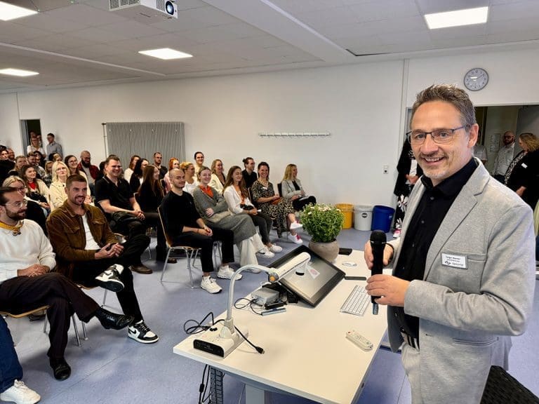 Holger Werner steht mit Mikrofon an einem Rednerpult vor einem Publikum in einem Seminarraum. Im Hintergrund sitzen zahlreiche Zuhörer und Zuhörerinnen und lächeln in Richtung Kamera.