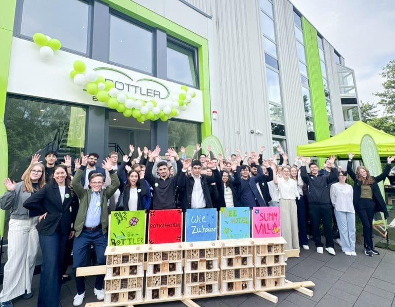 Neue Auszubildende vor dem Firmengebäude in Neheim, winkend, mit selbstgebauten Insektenhotels im Vordergrund.