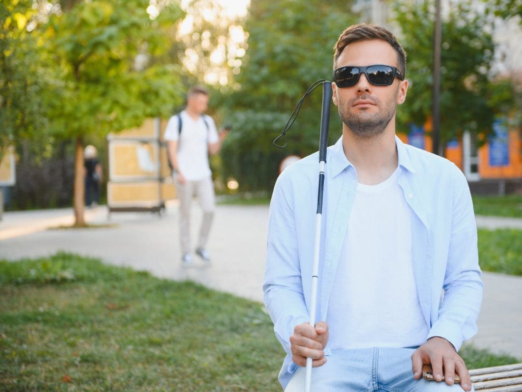 Ein junger Mann mit Blindenstock sitzt auf einer Parkbank in einem grünen Stadtpark.
