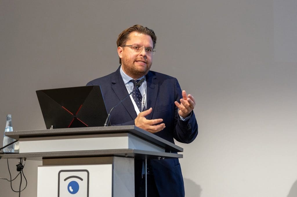Ein Redner im Anzug spricht am Podium und gestikuliert während einer Präsentation.